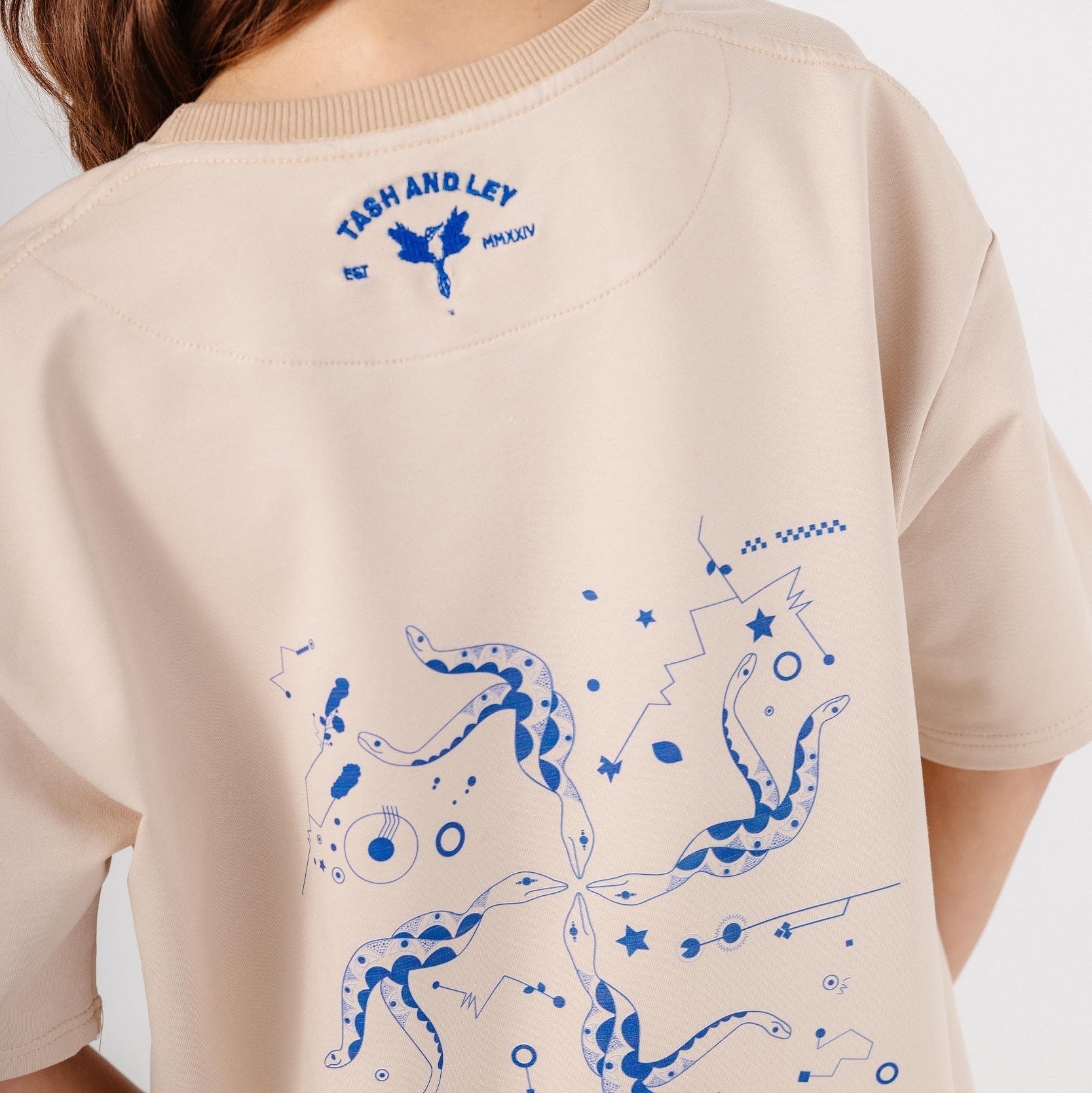 Model wearing beige top with blue embroidery and print design on back