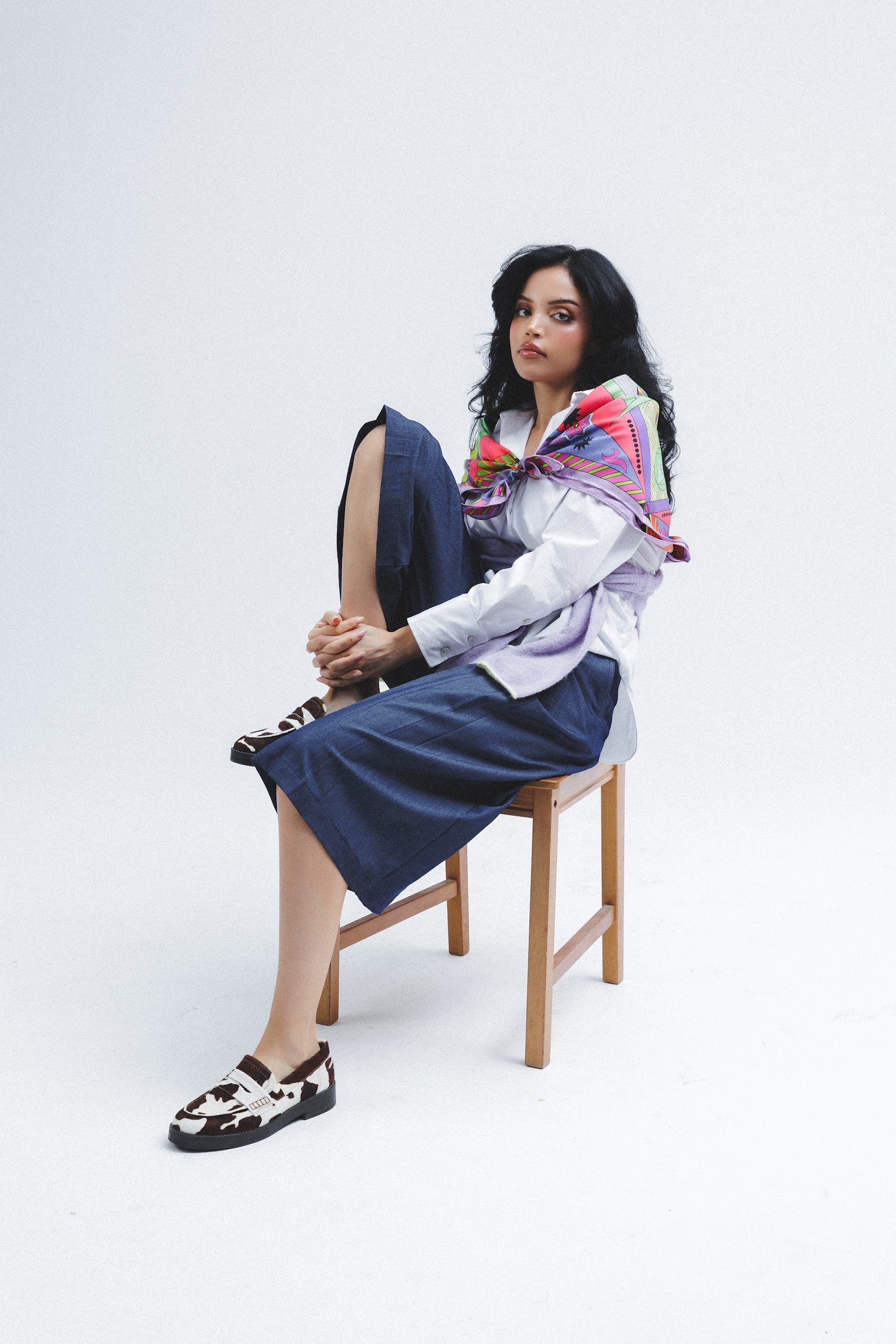 Model sitting on a chair wearing a white blouse, blue bermuda, and colorful scarf on her shoulders