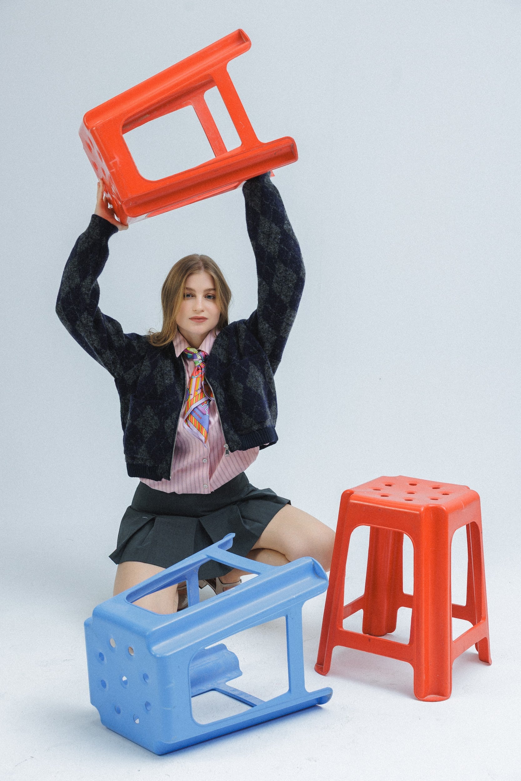 Model holding a red stool with two other stools on the floor wearing silk scarf as tie