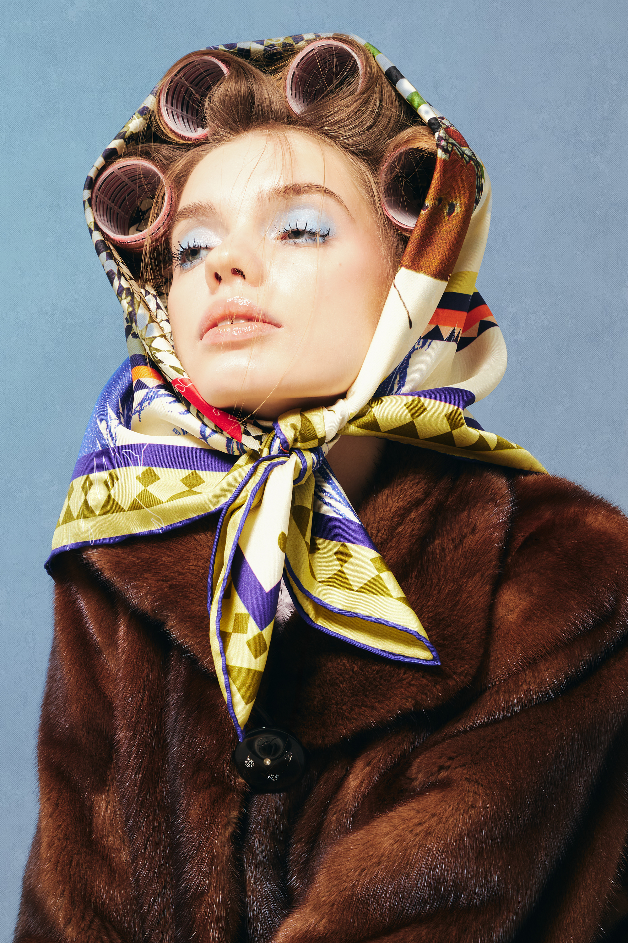 Woman wearing a patterned silk twill square scarf as a headscarf and brown fur coat against a blue background
