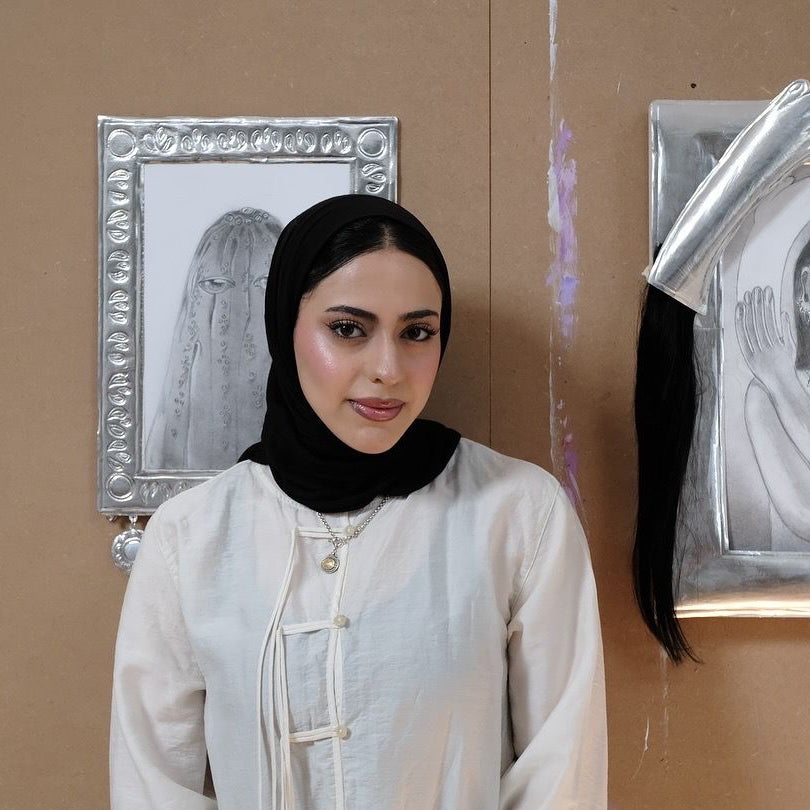 Artist Zainab Al Shibani standing in front of wall art with silver decorations and framed images.