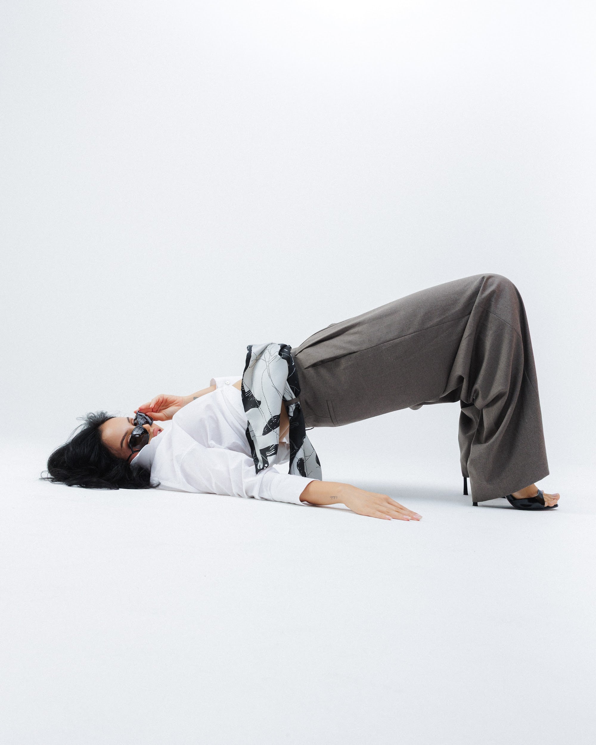 Model lying on the floor wearing a white shirt and gray pants with a silk scarf on the belt of her trousers