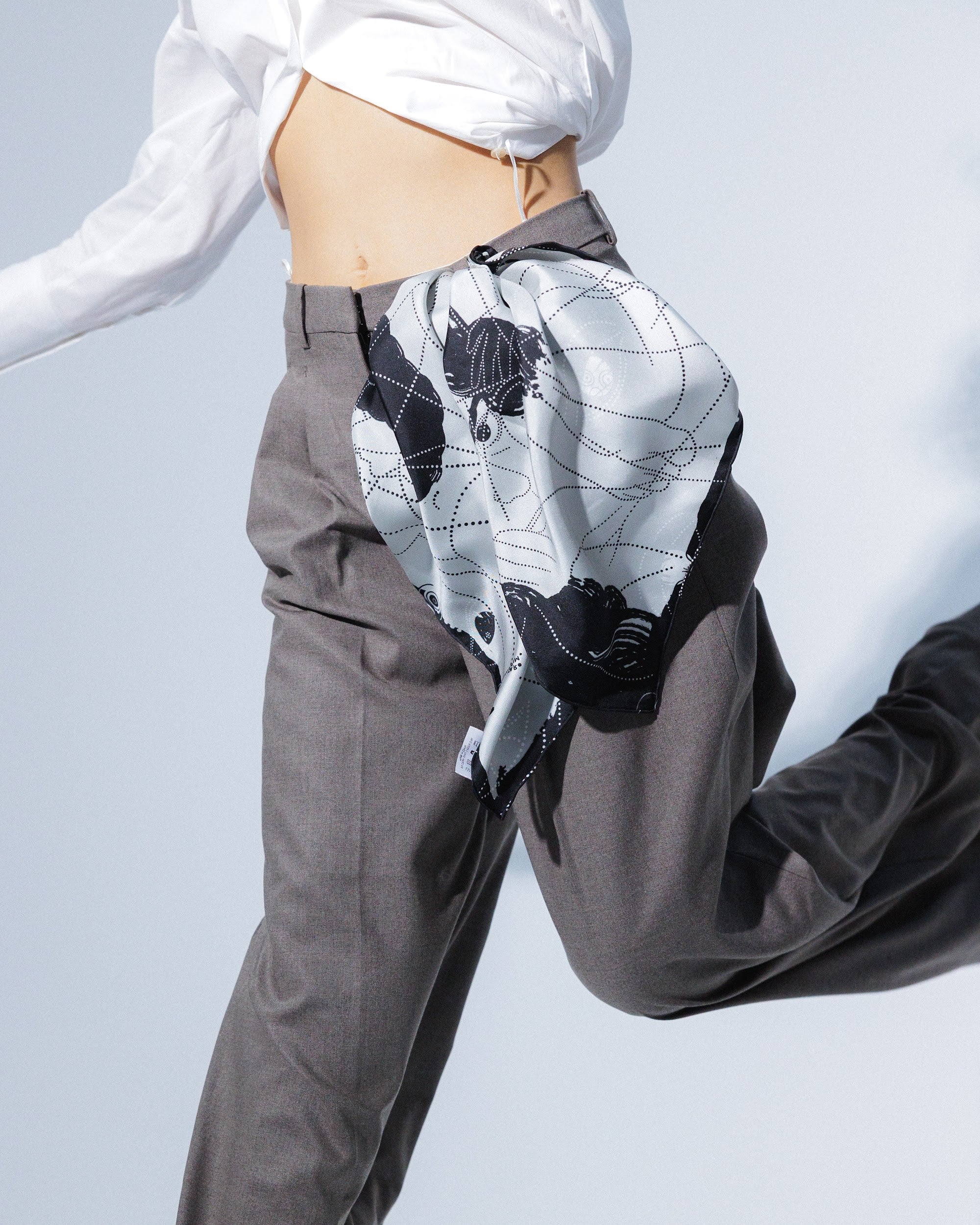 Model wearing gray pants and a white shirt with a silk scarf tied around her waist