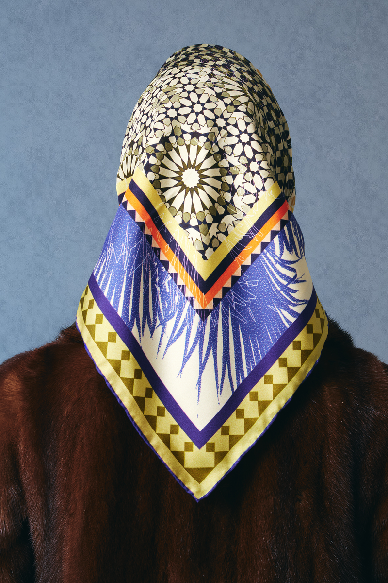 Person wearing a beige and blue silk twill square scarf as a headscarf with geometric patterns on a plain background